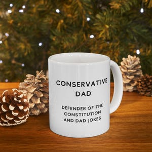 May include: White ceramic mug with the text "CONSERVATIVE DAD" above "DEFENDER OF THE CONSTITUTION AND DAD JOKES" in black. The mug has a curved handle and is surrounded by pinecones and a blurred green background.