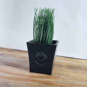 May include: A black square planter with a hidden camera, filled with green artificial grass. The planter is on a wooden surface. The text "DECALDINO.COM" is visible on the side of the planter.