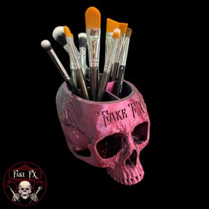 Puede incluir: Un portacepillos de maquillaje de color rosa en forma de calavera con las palabras "Fake FX" grabadas en la parte delantera. El portacepillos tiene dos compartimentos y está lleno de varios pinceles de maquillaje.