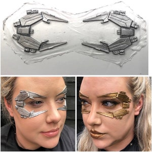 May include: A close-up of two silver and gold face makeup prosthetics. The prosthetics are shaped like a futuristic design with sharp angles and lines. The prosthetics are on a white background.