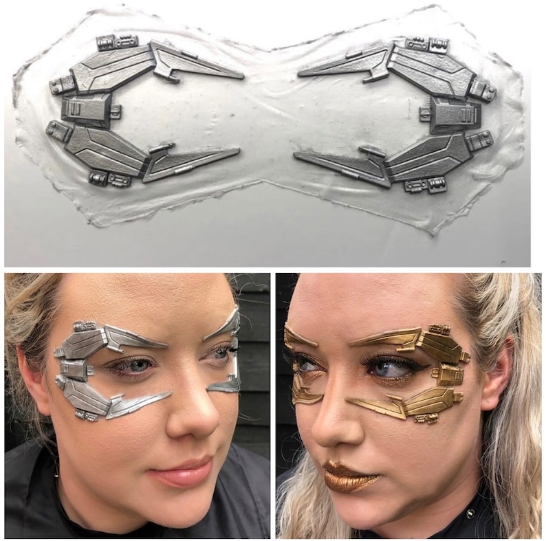 May include: A close-up of two silver and gold face makeup prosthetics. The prosthetics are shaped like a futuristic design with sharp angles and lines. The prosthetics are on a white background.