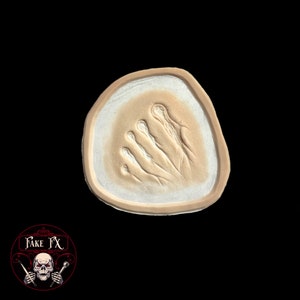 May include: A white silicone mold with a raised impression of a human hand. The mold is shaped like a rounded triangle and has a light brown border.