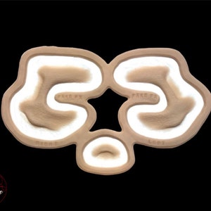 May include: A beige prosthetic makeup piece with a star-shaped central design and three curved sections. The text "FAKE FX", "RIGHT", and "LEFT" are visible. The piece has white and brown accents, set against a black background.