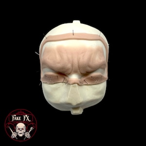 Peut inclure: Gros plan d'un moule de visage en mousse blanche avec un front texturé et des orbites. Le moule est piqué avec de petites épingles noires.