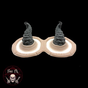 Puede incluir: Un par de cuernos protésicos para cosplay o maquillaje de efectos especiales. Los cuernos están hechos de un material flexible y están diseñados para ser fijados a la frente. Los cuernos son grises y tienen una superficie texturizada. La imagen muestra los cuernos en un molde con las palabras "Derecha" e "Izquierda" etiquetadas en cada lado.