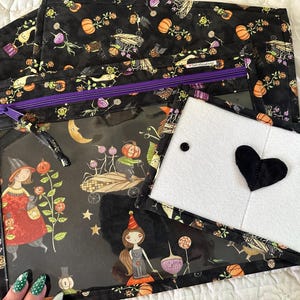 May include: A black fabric pouch with a clear window featuring a Halloween-themed print of pumpkins, witches, and stars. The pouch has a purple zipper and a separate white felt flap attached with a black heart-shaped accent and black snaps.