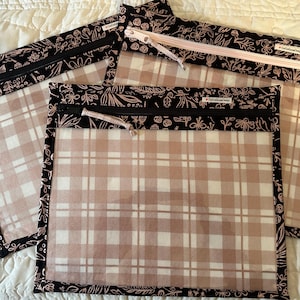May include: Three square zippered pouches featuring a clear center panel with a beige and white plaid pattern. The borders are black fabric with a white line-art floral print. Each pouch has a zipper closure with a fabric pull tab.