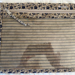 May include: Clear, rectangular pouch with a beige and blue striped pattern. The top edge features a floral print border and a zipper closure. The pouch is trimmed with a floral patterned fabric.