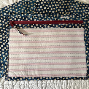 May include: A rectangular fabric pouch featuring a dark blue exterior with a repeating pattern of small cream-colored stars. The front panel displays a white background with thin horizontal red and blue stripes and a red zipper closure.
