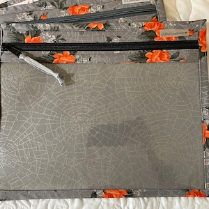 Peut inclure: Une pochette rectangulaire à fermeture éclair avec un centre en vinyle transparent orné d'un motif de toile d'araignée grise. La bordure est en tissu gris à motif floral orange. Une fermeture noire est sur le dessus avec l'étiquette DotDotGooseDesign.