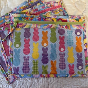 May include: Several colorful zippered pouches with a clear front panel. The front panel features a pattern of pastel-colored Easter bunny silhouettes on a light blue background. The zipper is green, and the edges are trimmed with a vibrant, psychedelic pattern.