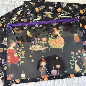 May include: A rectangular zippered pouch with a clear vinyl front panel and a black fabric border featuring a colorful Halloween-themed print of witches, pumpkins, owls, and stars. The pouch has a purple zipper closure and a black fabric pull tab.