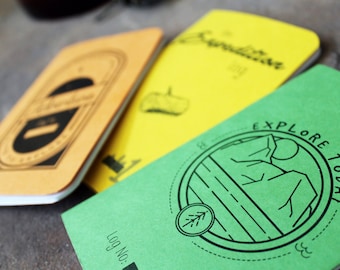 May include: Three small notebooks in yellow, orange, and green. The orange notebook has the word "Adventure" in a black, stylized font. The yellow notebook says "Expedition". The green notebook says "Explore Today" with a mountain graphic.