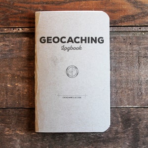 May include: A gray, rectangular logbook with the text "GEOCACHING Logbook" printed in black. A compass symbol is printed in the center of the book.