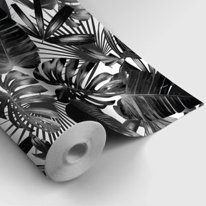 May include: A roll of wallpaper featuring a black and white tropical leaf pattern. The design includes large monstera leaves, palm fronds, and other foliage against a white background. The wallpaper is partially unrolled, revealing the paper's core.