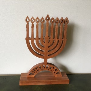 Puede incluir: Un menorá de madera con nueve candelabros, cada uno con forma de llama. La base presenta la palabra "Shalom" recortada. El menorá está hecho de madera de color marrón rojizo y se apoya sobre una base rectangular.