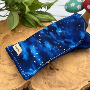 Puede incluir: Un estuche para gafas de galaxia azul y blanco con una etiqueta dorada que dice "Amelia Nova".