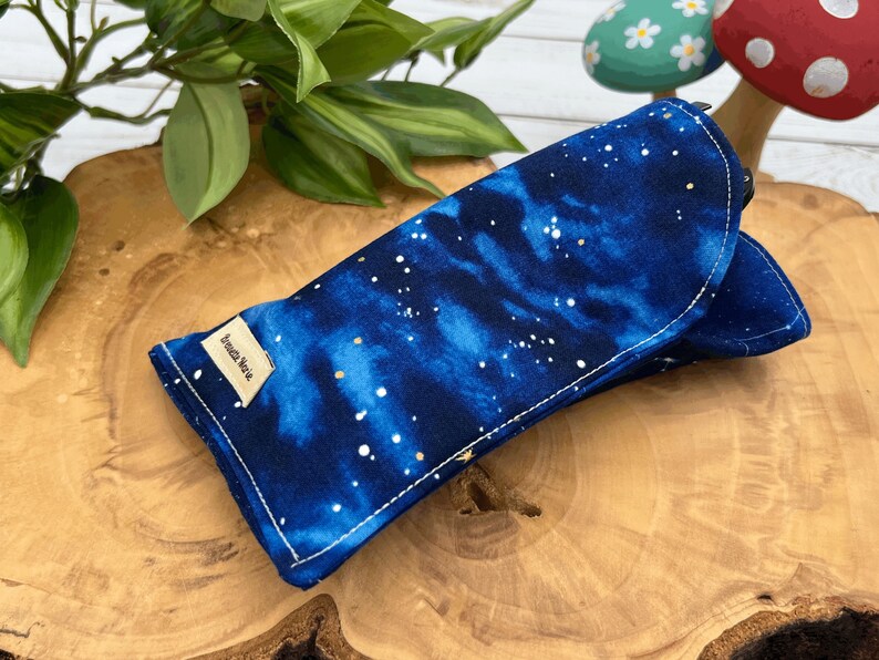 Puede incluir: Un estuche para gafas de galaxia azul y blanco con una etiqueta dorada que dice "Amelia Nova".