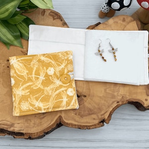 May include: A small yellow fabric pouch with a white dragonfly pattern and a button closure. The pouch is open and a pair of silver dragonfly earrings with brown beads are inside.