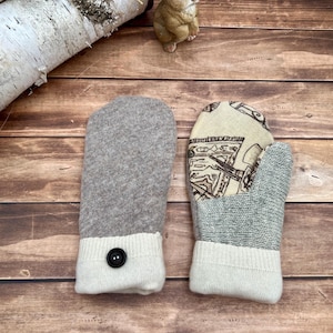 May include: A pair of mittens featuring a solid light brown knit fabric on one side and a cream-colored cuff with a single black button. The other side displays a patterned cream fabric with black line drawings and a gray knit section.