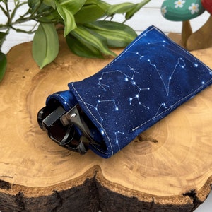 Puede incluir: Un estuche para gafas de tela azul con un patr&oacute;n de constelaci&oacute;n blanca. El estuche est&aacute; doblado y tiene unas gafas de sol dentro.