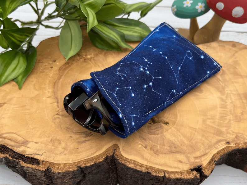 Puede incluir: Un estuche para gafas de tela azul con un patr&oacute;n de constelaci&oacute;n blanca. El estuche est&aacute; doblado y tiene unas gafas de sol dentro.