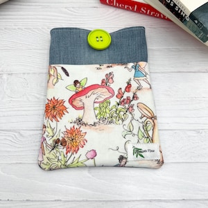 Puede incluir: Una funda de libro de tela con una parte superior de mezclilla y un cierre de botón. La tela principal presenta un diseño de cuento de hadas caprichoso con hadas, setas y flores en tonos naranja, rosa y verde. La etiqueta dice "Fiannette Marie".