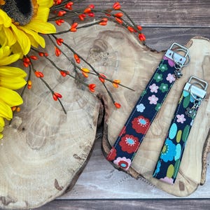 May include: Two fabric key fobs with floral patterns in red, pink, and blue on a navy background. The key fobs have silver hardware and a small flower charm. The fobs are on a wooden surface with sunflowers and orange berries.