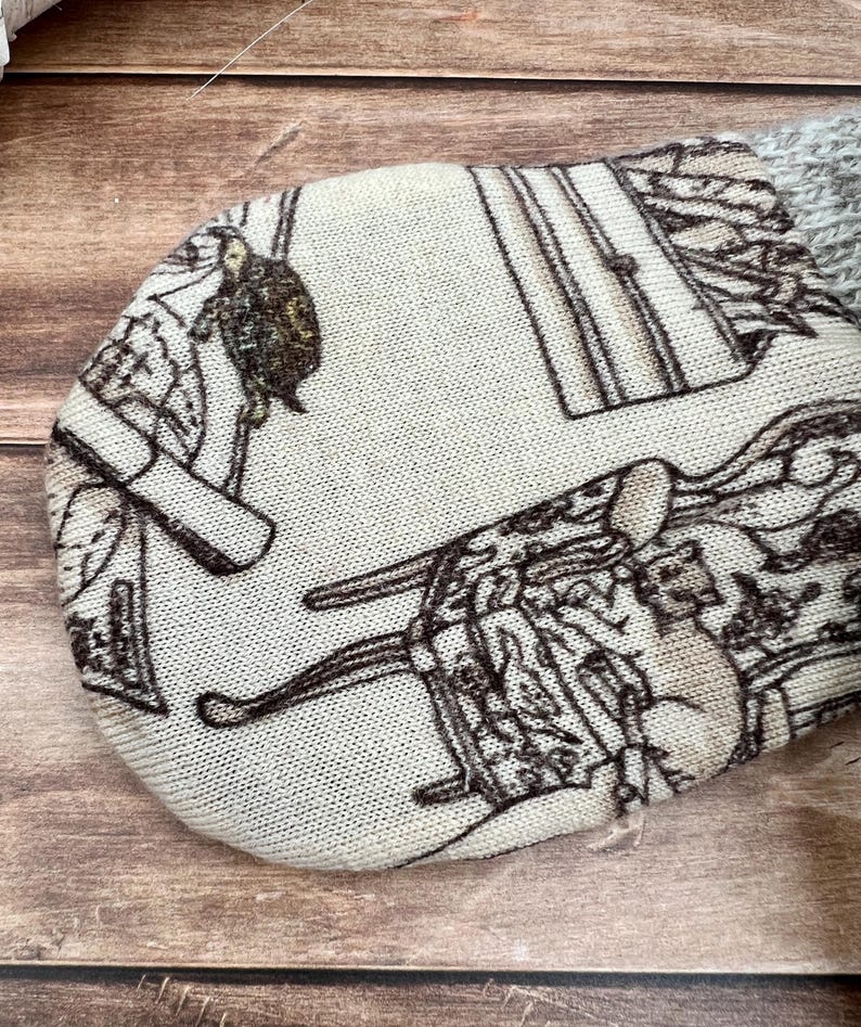 May include: A rounded fabric item featuring a cream-colored base with a dark brown printed pattern depicting various furniture pieces, including a chest of drawers and a chair, with a textured gray knit fabric attached at the top edge.