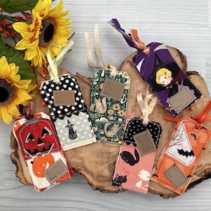 May include: A selection of handmade Halloween gift tags. Designs feature a pumpkin, witch, black cat, and bats. Each tag includes a small label for personalisation and is decorated with ribbons.