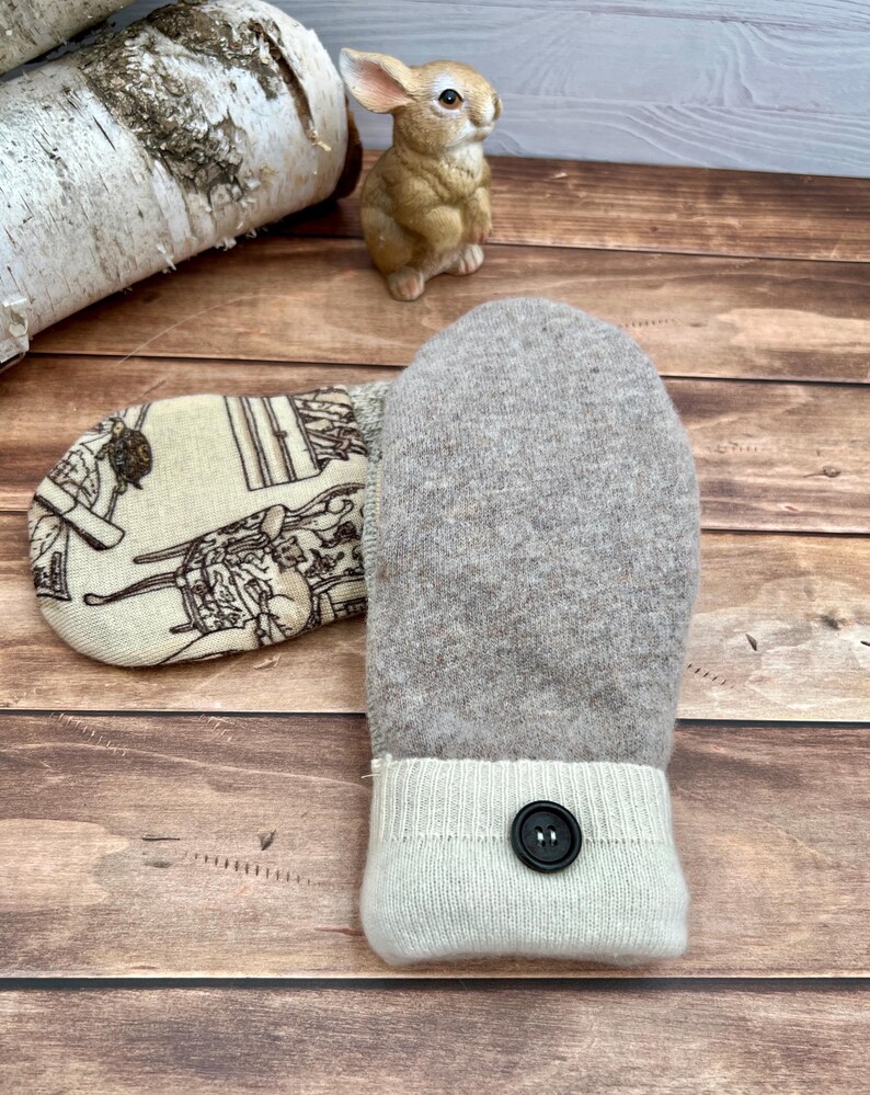 May include: A pair of mittens featuring one solid light brown felted wool side and one cream-colored side with a black line-drawn pattern of furniture and cats. Each mitten has a ribbed cream cuff accented with a single black button.