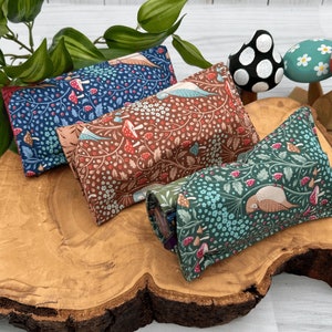 May include: Three fabric pouches with floral and bird patterns. The pouches are arranged on a wooden slice. The pouches are in shades of blue, brown, and green.