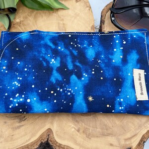 Puede incluir: Un estuche para gafas de tela azul y negro con un patr&oacute;n de cielo estrellado blanco y dorado. El estuche tiene una peque&ntilde;a etiqueta blanca con el texto "Brewette Marie" en &eacute;l.