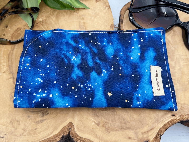 Puede incluir: Un estuche para gafas de tela azul y negro con un patr&oacute;n de cielo estrellado blanco y dorado. El estuche tiene una peque&ntilde;a etiqueta blanca con el texto "Brewette Marie" en &eacute;l.