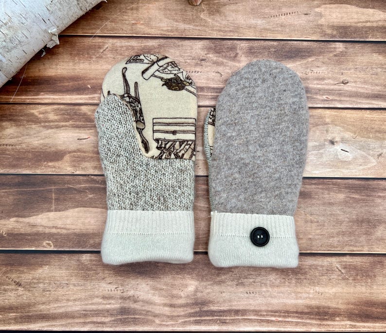 May include: A pair of mittens featuring a cream-colored fabric with a brown line-art pattern on one side and solid heathered gray wool on the other. The cuffs are made of ribbed cream knit fabric, with a single black button on one cuff.