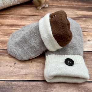 May include: A pair of gray mittens featuring a dark brown fleece lining and a cream-colored ribbed cuff. One mitten is accented with a single black button on the cuff. The mittens are constructed from a soft, knit fabric with a folded cuff design.