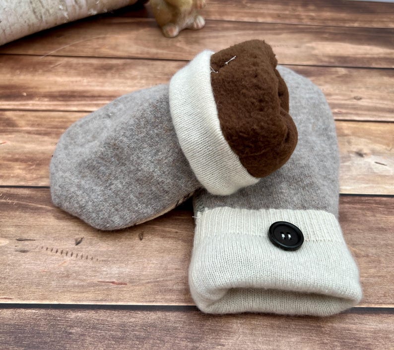 May include: A pair of gray mittens featuring a dark brown fleece lining and a cream-colored ribbed cuff. One mitten is accented with a single black button on the cuff. The mittens are constructed from a soft, knit fabric with a folded cuff design.