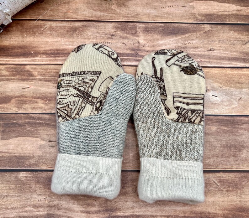 May include: A pair of mittens featuring a cream-colored fabric upper with a brown line-art pattern of various objects. The lower section is made of a textured gray knit fabric, finished with a solid cream-colored ribbed knit cuff.