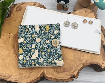 May include: A teal blue fabric jewelry box with a floral and cat print. The box has a button closure and is open to reveal a white fabric interior with two pairs of flower-shaped earrings and two small round earrings.