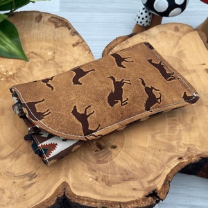 Op de afbeelding: Een bruine leren portemonnee met een herhaald patroon van donkerbruine rodeo figuren. De portemonnee is open en toont een patroon binnenkant met een Native American design in rood, wit en bruin. Het rust op een houten oppervlak.