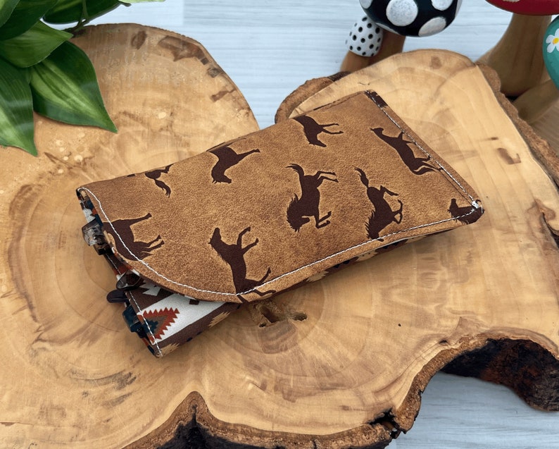Op de afbeelding: Een bruine leren portemonnee met een herhaald patroon van donkerbruine rodeo figuren. De portemonnee is open en toont een patroon binnenkant met een Native American design in rood, wit en bruin. Het rust op een houten oppervlak.