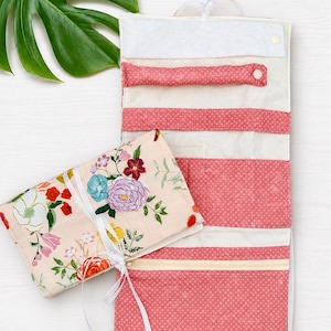 Op de afbeelding: Een stoffen organizer om op te rollen met een lichtroze bloemenprint en witte linten. De binnenkant toont afwisselende secties van effen crèmekleurige stof en roze stof met witte stippen, inclusief een ritsvak en drukknopen.