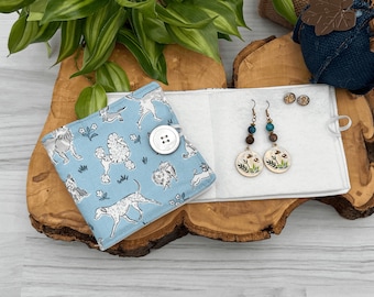 May include: A blue fabric jewelry organizer with a button closure. The organizer is decorated with a pattern of white dogs. The organizer is open to reveal a white velvet interior with two pairs of earrings. One pair of earrings features a brown wooden bead and a turquoise stone. The other pair of earrings features a round, white ceramic charm with a floral design.