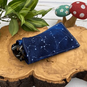 Puede incluir: Una funda para gafas de sol de tela azul con constelaciones blancas impresas en ella. La funda est&aacute; sobre una superficie de madera con unas gafas de sol dentro.