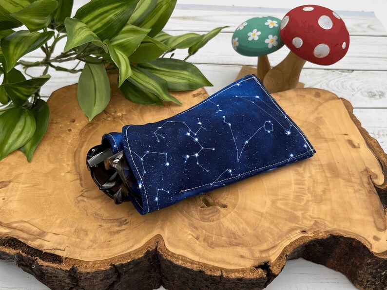 Puede incluir: Una funda para gafas de sol de tela azul con constelaciones blancas impresas en ella. La funda est&aacute; sobre una superficie de madera con unas gafas de sol dentro.