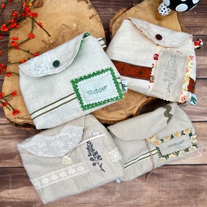 Puede incluir: Cuatro bolsas de tela beige con cierres de solapa, cada una con adornos únicos. Los detalles incluyen encaje, diseños florales bordados y adornos decorativos. Las bolsas están dispuestas sobre una superficie de madera, mostrando una estética rústica.