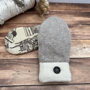 May include: A pair of mittens featuring a light gray wool-like texture on one side and a cream-colored fabric with a black line-art illustration of furniture and cats on the other. Each mitten has a cream ribbed cuff with a single black button.