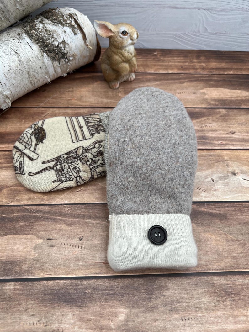 May include: A pair of mittens featuring a light gray wool-like texture on one side and a cream-colored fabric with a black line-art illustration of furniture and cats on the other. Each mitten has a cream ribbed cuff with a single black button.