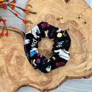 May include: Black scrunchie with a cat-themed pattern. The fabric features white cat silhouettes, the word "MEOW" in pink, and other colorful cat-related graphics. The scrunchie is displayed on a wooden surface with a sunflower and red berries.