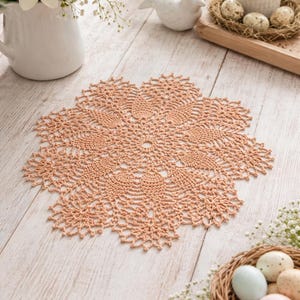 May include: A peach-colored, crocheted doily with a detailed floral pattern sits on a light wood surface. The doily is surrounded by Easter decorations, including eggs in a nest, flowers, and a candle.